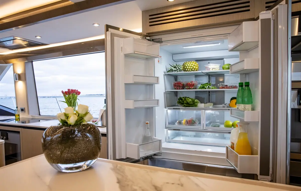 Open refrigerator filled with fresh produce, including pineapple, lettuce, and drinks, in a modern kitchen with a waterfront view.